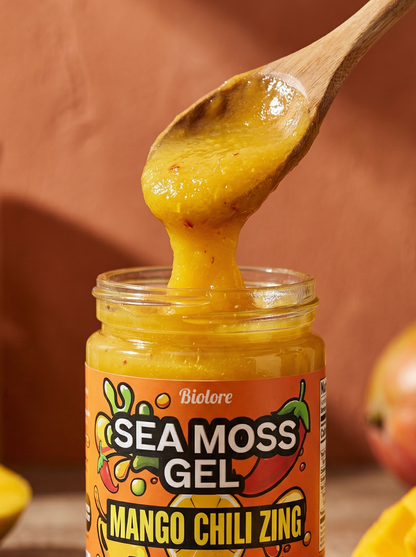 Jar of Sea Moss Gel with a wooden spoon lifting some out, surrounded by mangoes and chili peppers on a wooden surface.