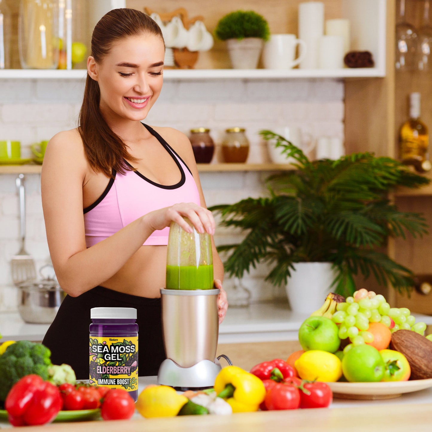 Woman preparing a green smoothie in a kitchen surrounded by fresh fruits with elderberry sea moss gel placed on the counter.